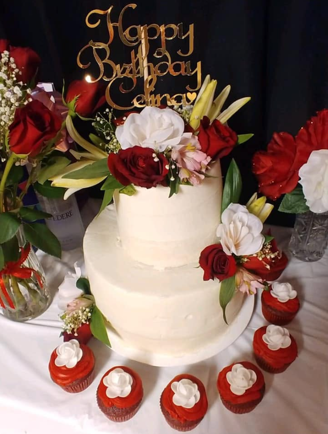Two tier celebration cake in white buttercream, dressed with red and white roses, lilies and matching red velvet cupcakes, by Bianca Mendoza